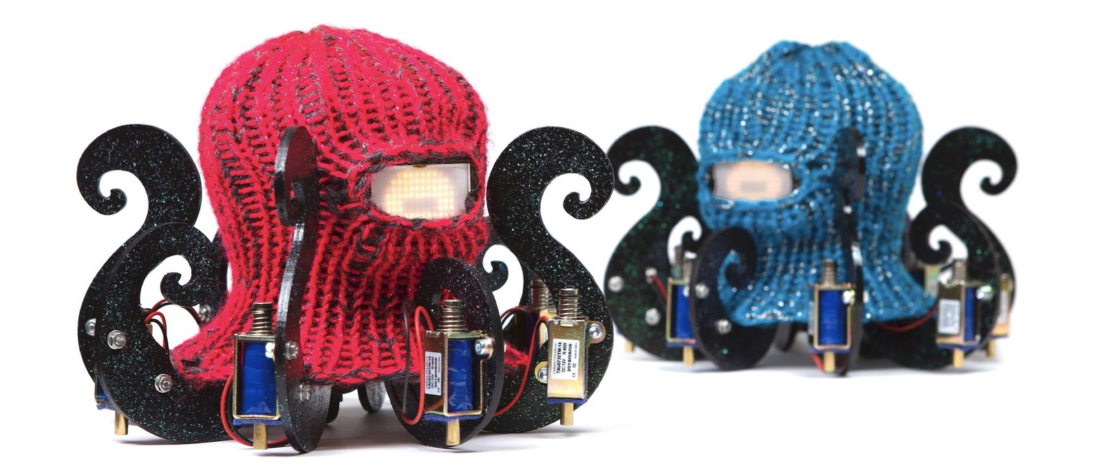 Two small robots shaped like octopuses, one with a red knitted body and one with a blue one, each with a glowing dot-matrix face and solenoids mounted on black tentacles.