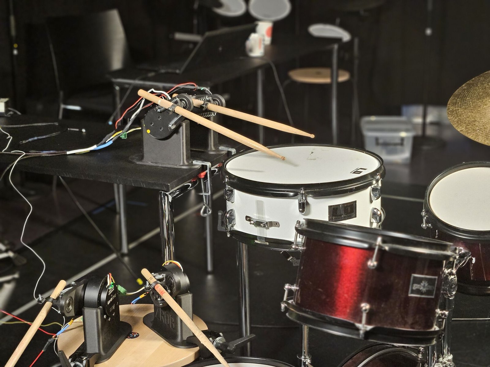 Two robotic arms holding drumsticks over a snare drum and a wooden practice pad in a dark laboratory, with springs, motors, and coloured wires visible.