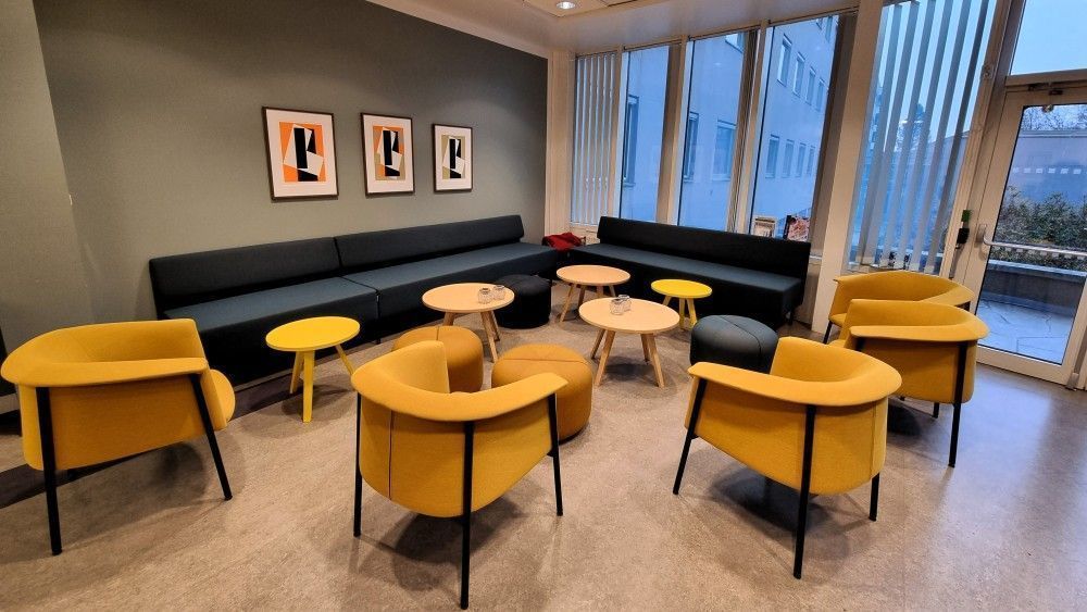 The common area: yellow armchairs, dark sofas, low round tables and abstract prints on a grey wall, by tall windows